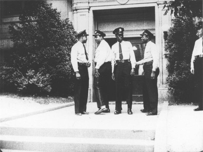 Four black police officers stand together outside. Another officer stands off to the side. 
