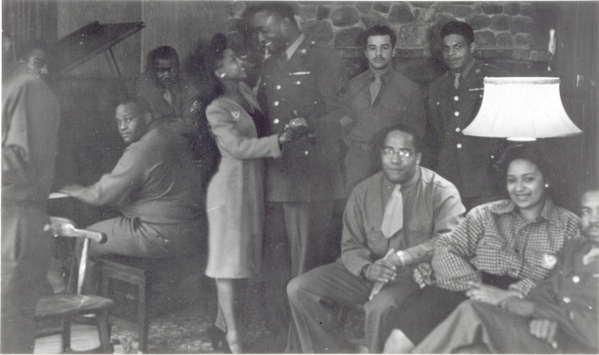 A large indoor gathering of black men and women. The men are wearing uniforms. One person is seated at a piano, a couple dances, and numerous other people are standing and seated around them. 