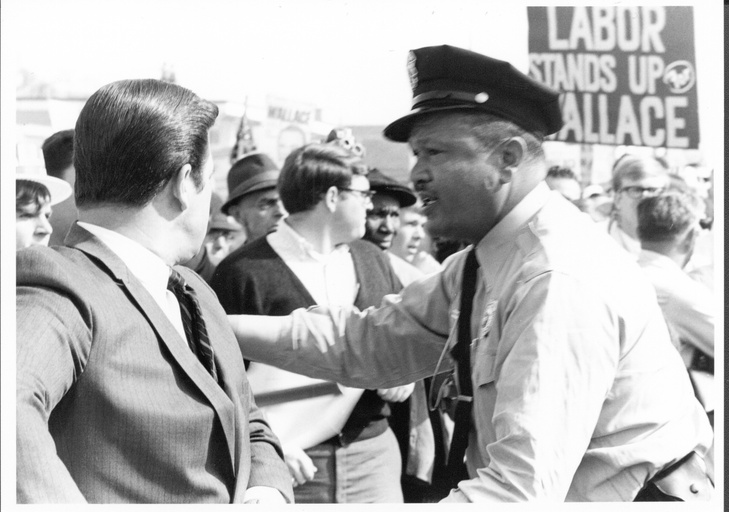 An African-American police officer works on crowd control at the Wallace-for-president rally at the Durham police headquarters. A protest sign in the background says "Labor Stands Up for Wallace."