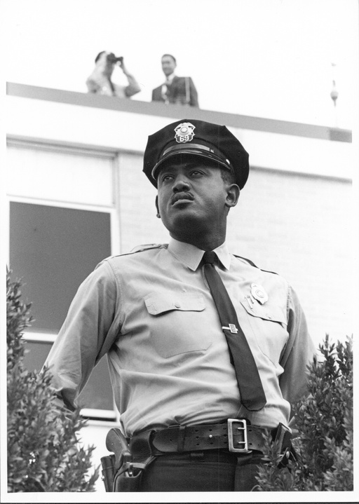 An African-American police officer. In the background two people are standing on a roof. 