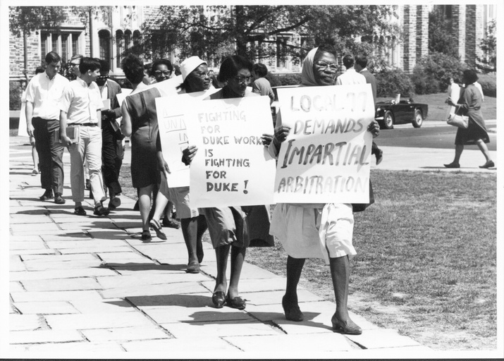 A line of protesters with signs marching on Duke's campus. In the foreground are African-American women and in the background are some white male students. 