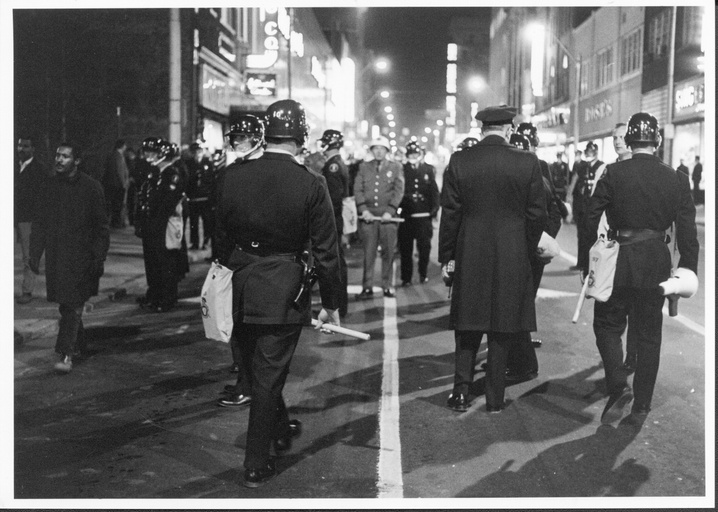 A large group of people in the street. Most people in the image are police in riot gear with their backs to the camera. 