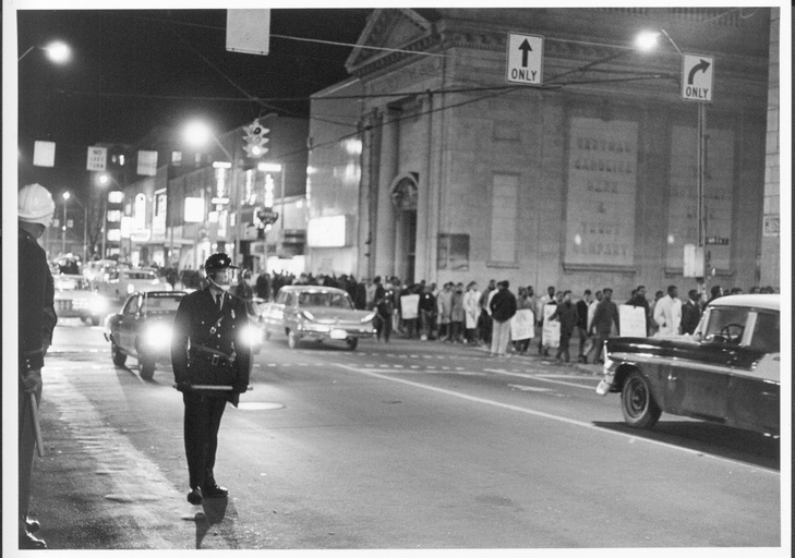 A police officer directing traffic with a large group of protesters in the background.