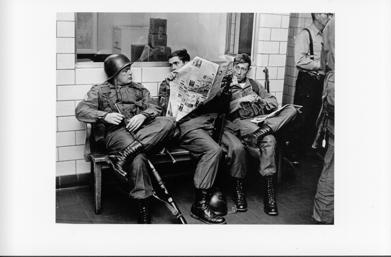 Three white, armed National Guard soldiers seated on a bench. One of them is reading a newspaper. 
