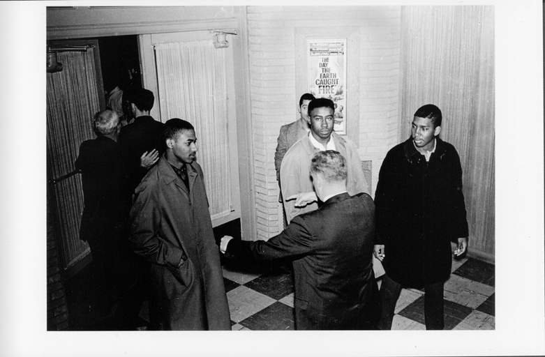 Four black men inside a lobby facing a white man with his back to the camera. Other people are visible in the background. 