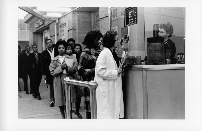 A black woman stands at a ticket counter speaking to a white woman ticket seller. A long line of black people is formed behind her. 