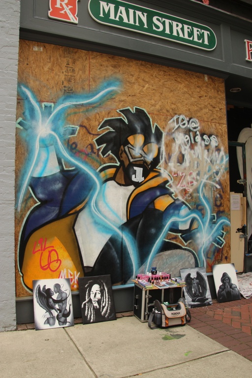 A spray painted black man in the style of a comic book superhero. It is made to look like electricity is coming out of his hands. On the sidewalk in front of the mural are vinyl album covers of black musicians. 