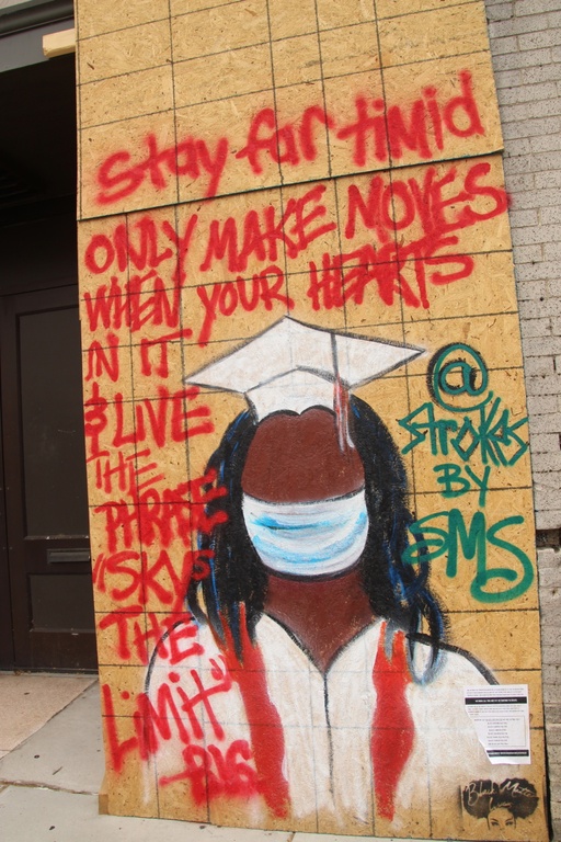 A mural depicting a black woman in a white graduation cap and gown wearing a mask over her face. Lyrics by rapper Notorious BIG are shown: "stay far timid only make moves when your hearts in it & live the phrase 'skys the limit.'" 