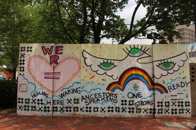 A large white mural with hearts, clouds, eyes, and a rainbow and the words "healing here waking ancestors speaking one strong ready."