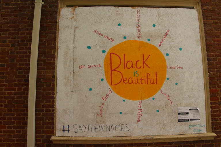 An image made to look like the sun with a yellow circle containing the words "black is beautiful." The names of black people murdered by police point out from the circle like rays of light. The hashtag #saytheirnames appears at the bottom of the mural.