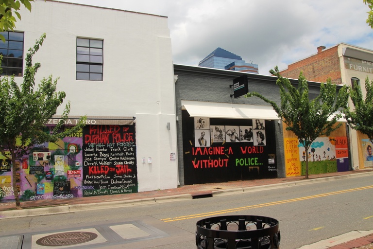 A wide shot showing multiple murals on storefronts and windows. Some of the words shown are "Killed by Durham Police," "Killed in Jail," "Imagine a World Without Police," "Every little thing is gonna be alright," and "Black as Eva."
