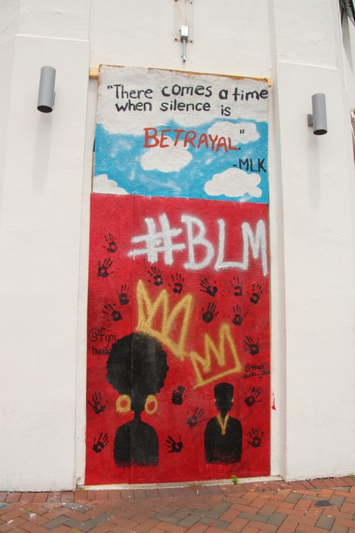 A red mural with the silhouette of a black man and woman with crowns, handprints all around them, and the hashtag #blm. Above that is a blue sky with clouds and the words "There comes a time when silence is betrayal - MLK."
