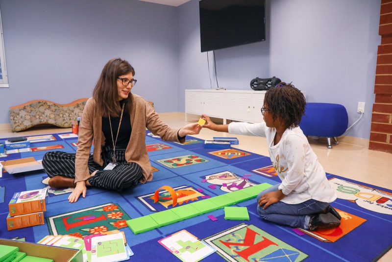 A child hands a coding toy to a librarian while sitting on a large colorful rug