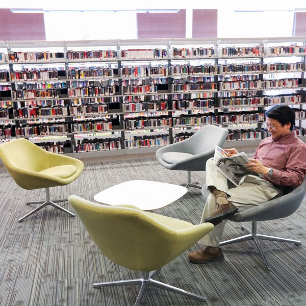 A man sits in a chair in front of rows of bookshelves and reads a newspaper
