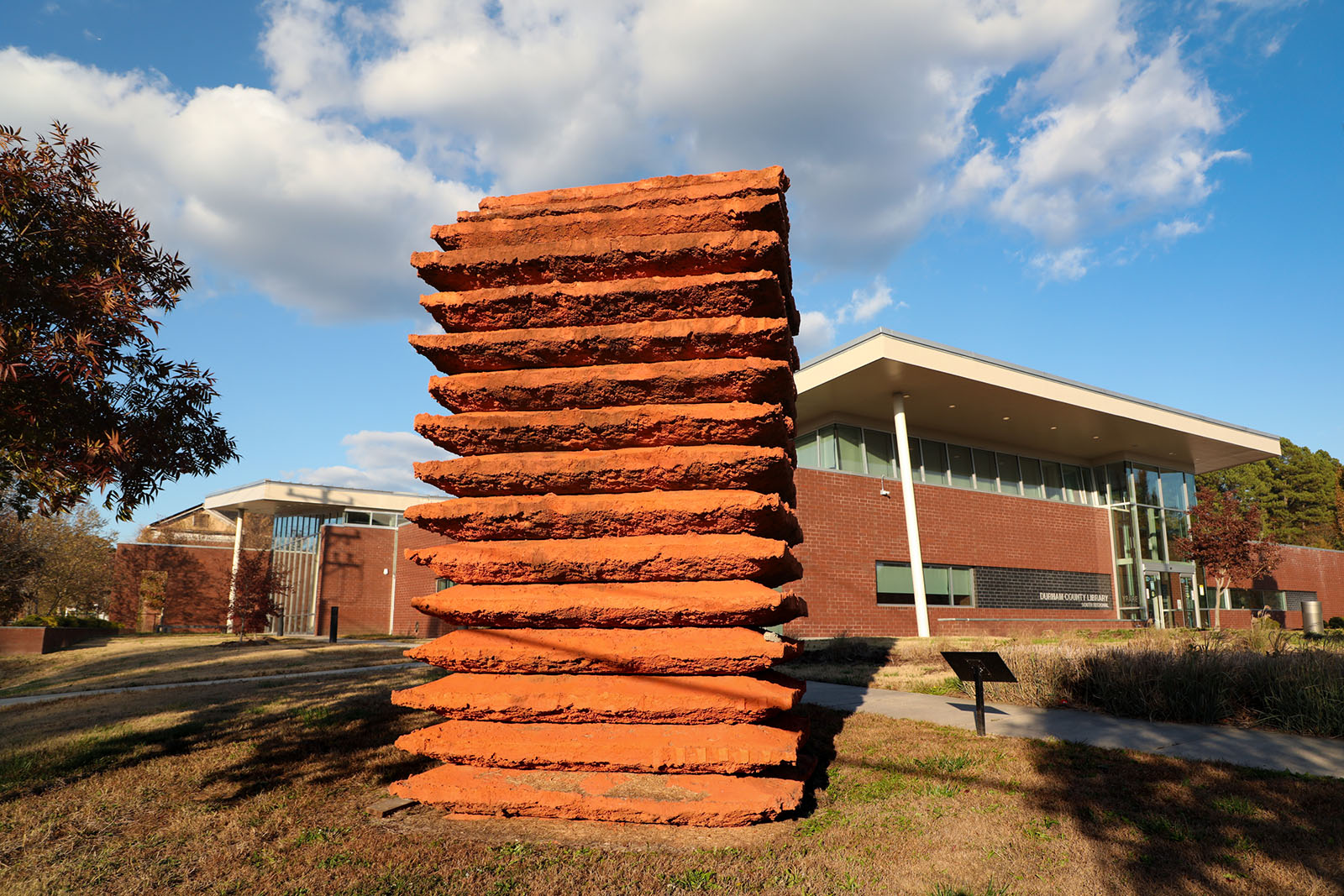 A tall, earth-colored sculpture, in the shape of a stack of slabs, standing in front of a building