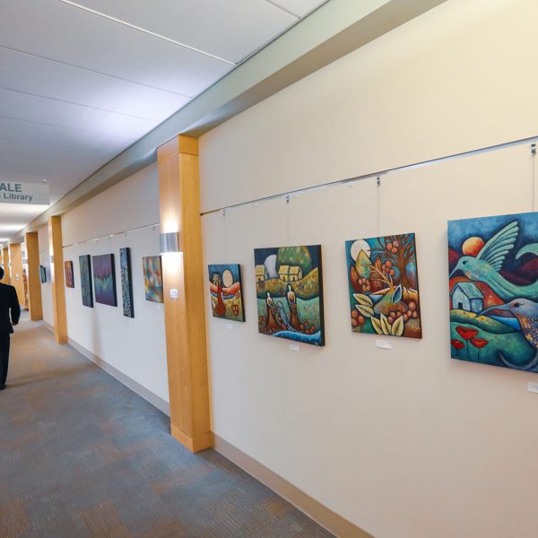 Colorful paintings with animals and landscapes hang in a line along a wall. A person walks along a corridor alongside