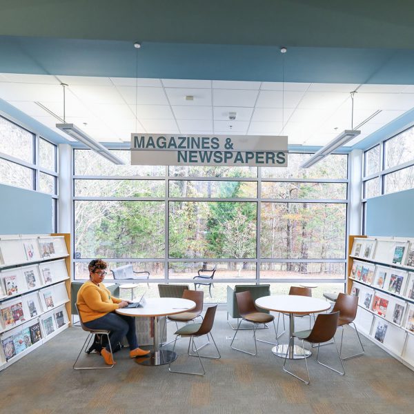 Brightly lit alcove with a sign overhead saying "Magazines & Newspapers", shelves of magazines along the side, and a wall of windows in the back. There are two round tables in the center with a person sitting at one working on a laptop