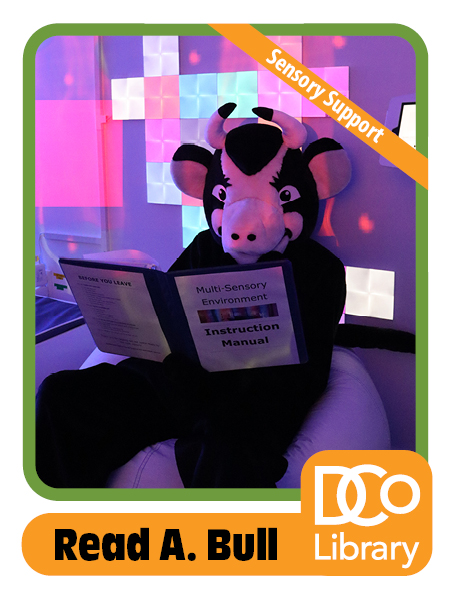 Costumed mascot Read A. Bull sitting on a beanbag chair in a space with gentle purple lighting, reading from a binder that says "multi-sensory environment instruction manual"