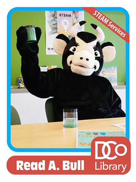 Costumed mascot Read A. Bull sitting at a work table and holding up a jar of a sciencey-looking substance