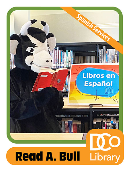 Costumed mascot Read A. Bull reading a book next to a bookshelf with a sign on top saying "Libros en Español"