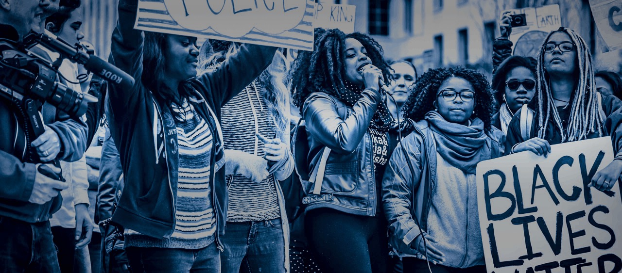 Group of young Black women speaking in front of news cameras at a protest, one holding a Black Lives Matter sign in the foreground