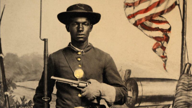 Civil War-era photograph: African American soldier in Union uniform with a rifle-musket and revolver in front of painted backdrop showing weapons and American flag