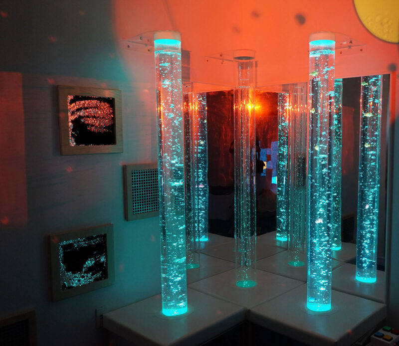 Tall tubes full of clear liquid with glowing blue bubbles throughout, in a corner of a room with low orange lighting and mirrors on the walls