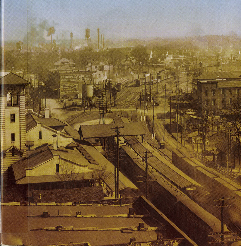 Sepia image of early Durham, with a train in the foreground and tracks extending toward the distance, wood and brick buildings, and lots of water towers and smokestacks on the horizon.